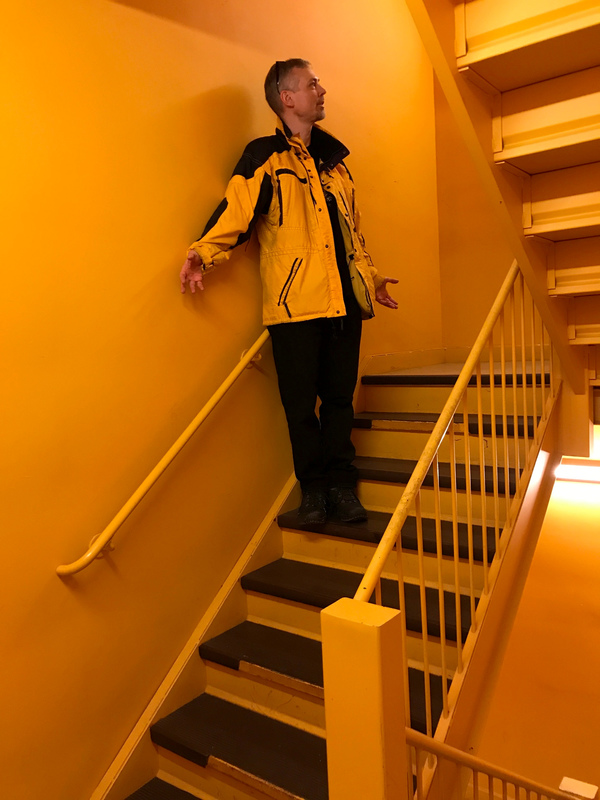 Stairwell with black steps and a yellow wall, me in black pants and a yellow and black jacket.