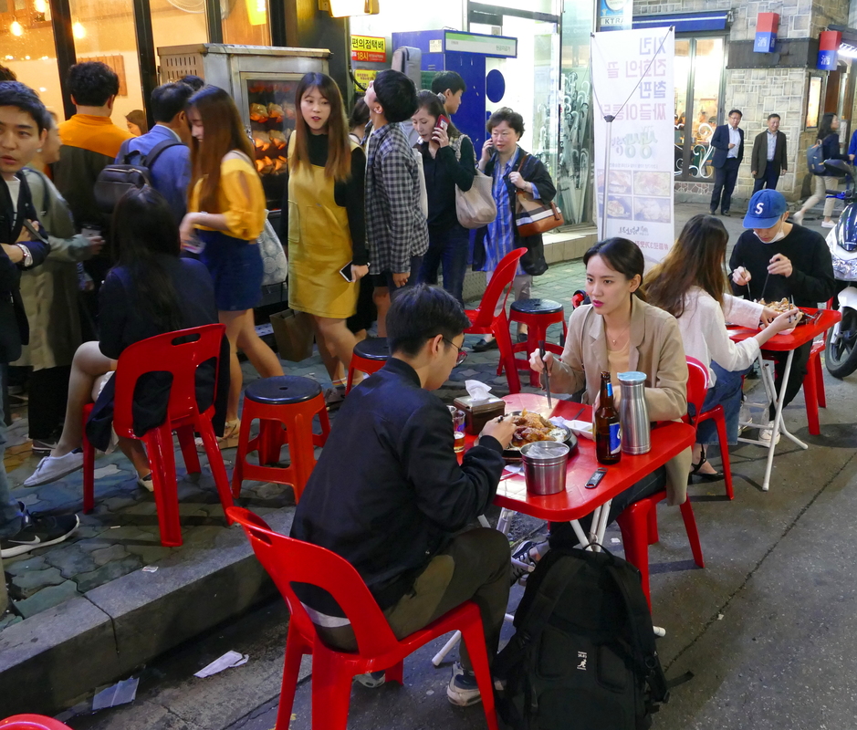 people on red plastic stools at small tables, eating on the street