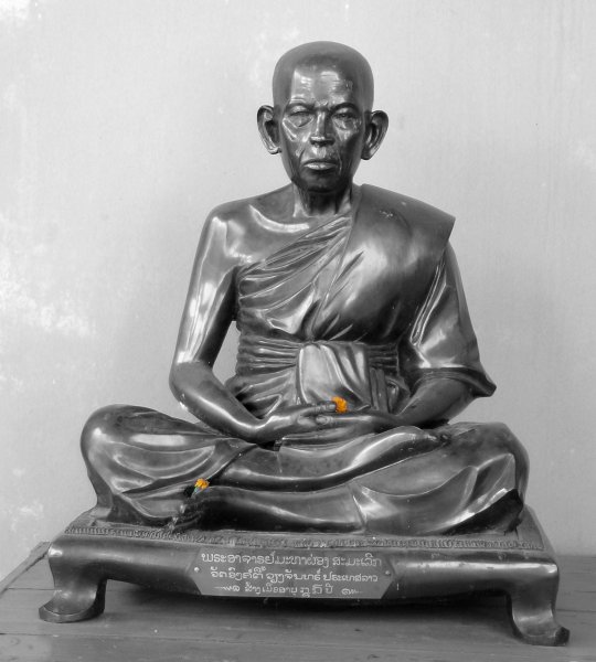 A sculpture of a monk sitting in half lotus with a couple small flowers placed on the sculpture.